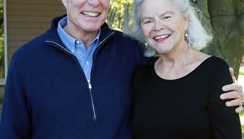 Gordon and Llura Gund are standing side by side smiling. Gordon is wearing a light blue dress shirt with a navy blue jacket over it. Llura is wearing a black long sleeved shirt.