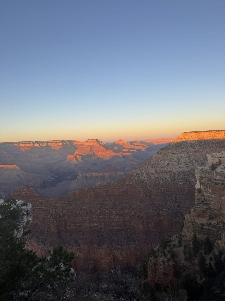 A photo of the Grand Canyon at sunset facing east. The sky is blue at the top with orange hues closest to the rocks. The top of the rocks are a vibrant red while the rest of the canyon is absorbed in shadows.