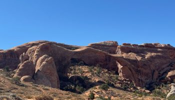 A wide desert landscape featuring a massive natural sandstone arch stretching between two rocky cliffs. The scene is filled with warm red and tan rock formations, scattered shrubs, and dry brush under a clear, bright blue sky.