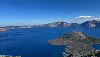 A sweeping view of Crater Lake on a bright, clear day, showing deep blue water surrounded by steep rocky cliffs and forested slopes. In the foreground, Wizard Island rises from the lake, cone-shaped and covered in pine trees. The sky above is vivid blue with a few scattered clouds.